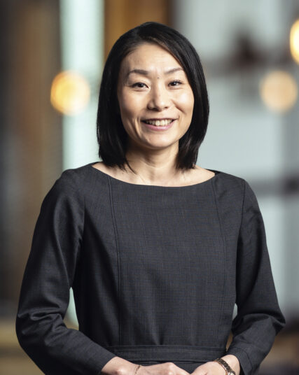 The image captures a woman with a gracious smile, short black hair, wearing a chic dark grey top. She stands with her hands gently clasped in front of her, against a backdrop featuring warm bokeh lights, suggesting a sophisticated indoor setting.