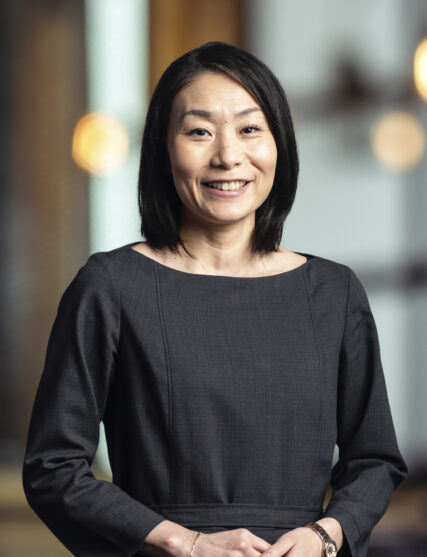 The image captures a woman with a gracious smile, short black hair, wearing a chic dark grey top. She stands with her hands gently clasped in front of her, against a backdrop featuring warm bokeh lights, suggesting a sophisticated indoor setting.