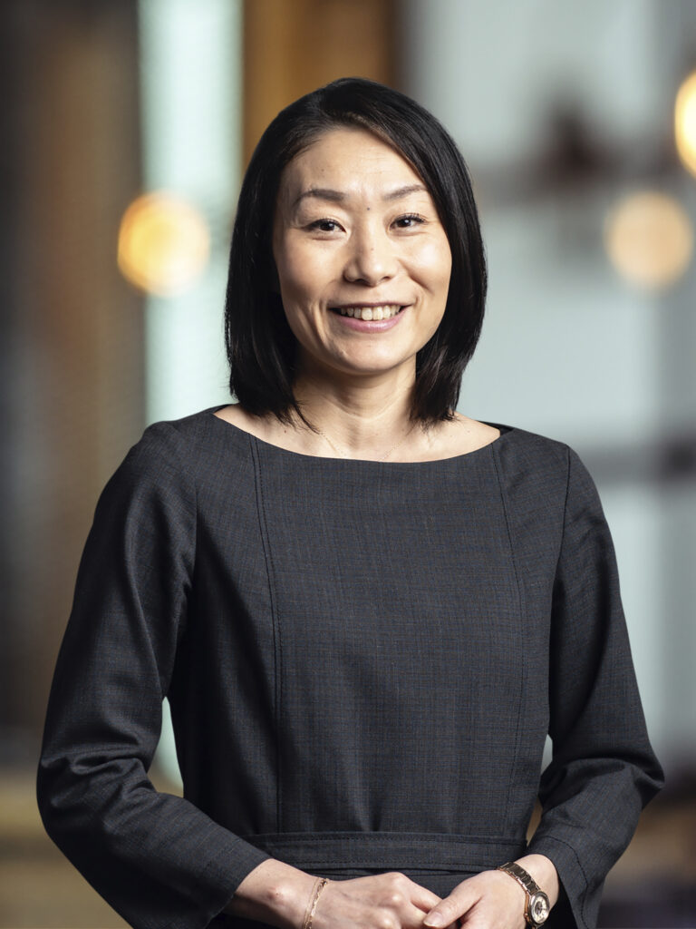 The image captures a woman with a gracious smile, short black hair, wearing a chic dark grey top. She stands with her hands gently clasped in front of her, against a backdrop featuring warm bokeh lights, suggesting a sophisticated indoor setting.