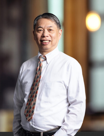 The photo captures a man with a friendly demeanor, wearing a crisp white shirt, a checkered tie in shades of orange and grey, and a belt. He stands with his hands casually in his pockets, against an out-of-focus background illuminated by warm light, indicative of an interior office space.
