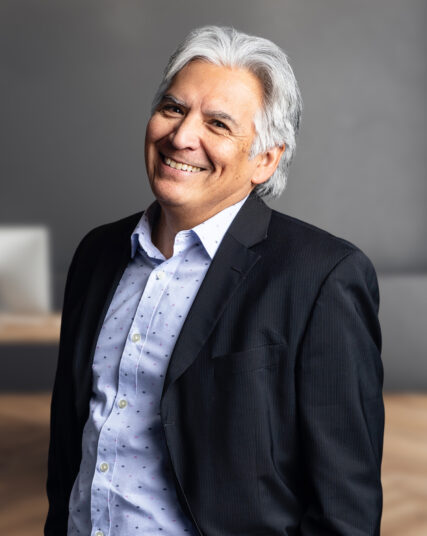 The image depicts a man with silver hair and a genuine smile, wearing a dark suit jacket over a light blue shirt with a subtle pattern. The neutral grey background is softly focused, highlighting the subject in a professional setting.