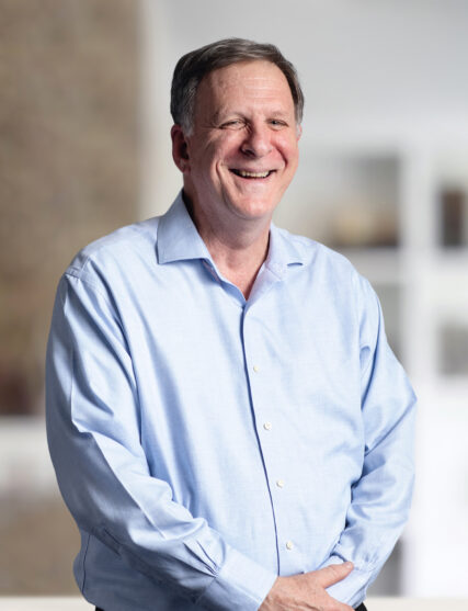 This photograph captures a man with a wide, sincere smile, wearing a light blue button-up shirt. He is standing with one hand in his pocket, against a backdrop that is light and indistinct, giving the impression of a relaxed yet professional setting.