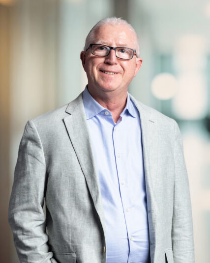 The image showcases a man with closely cropped white hair and glasses, wearing a light grey suit jacket and a pale blue shirt. He stands with a slight smile, hands in his pockets, against a background blurred with hints of green and beige, giving a calm and professional atmosphere.