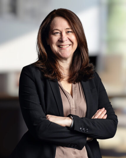 The image shows a woman with shoulder-length brown hair and a content smile, dressed in a professional black blazer and beige blouse. She stands with her arms crossed, conveying a confident and approachable stance against a softly blurred background suggestive of an office space.