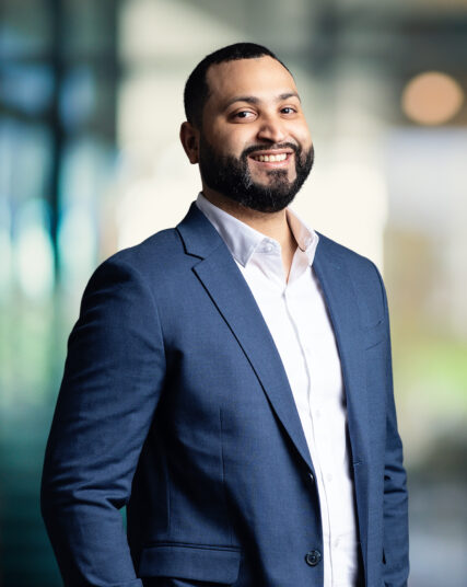 The image features a man with a beard and a pleasant smile, dressed in a navy blue suit with a white shirt. The background is light and airy with a soft focus, suggesting a spacious indoor setting with natural light streaming through.