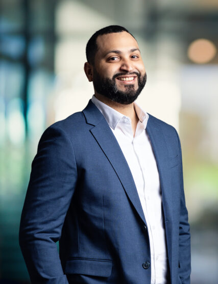 The image features a man with a beard and a pleasant smile, dressed in a navy blue suit with a white shirt. The background is light and airy with a soft focus, suggesting a spacious indoor setting with natural light streaming through.