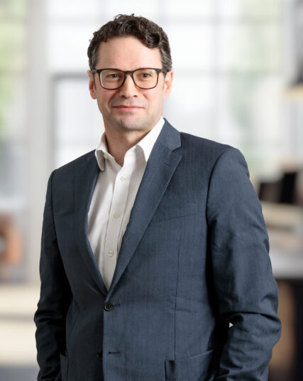 The image features a person in a dark jacket over a light shirt, standing indoors.