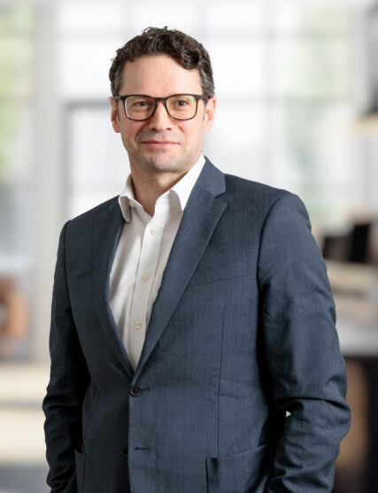 The image features a person in a dark jacket over a light shirt, standing indoors.