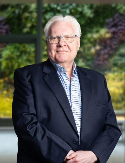 The image shows an elder man with white hair, wearing glasses, a navy blazer, and a light blue patterned shirt. He appears relaxed and confident, standing with one hand slightly clasping the other in front of him, against a blurred background of lush greenery.