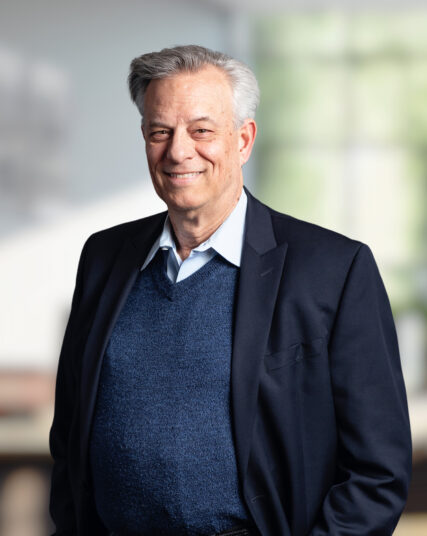 The image depicts a mature man with greying hair and a friendly smile, wearing a dark suit jacket over a blue sweater and a collared shirt. He stands with his arms slightly crossed, radiating confidence and professionalism against a blurred office background.