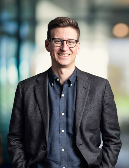 The image features a smiling man with glasses, short hair, and a smart-casual appearance, wearing a dark grey blazer over a navy shirt. The background has a bokeh effect with soft, circular light spots, suggesting a modern and dynamic office environment.