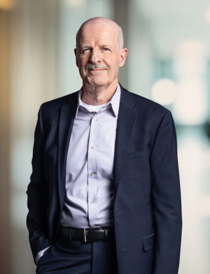 This image captures a distinguished looking man with a mustache, presenting a subtle smile and wearing a tailored dark suit with a light blue button-down shirt. He stands confidently, hands in pockets, against a background that is bright and blurred, likely an office setting with natural lighting.