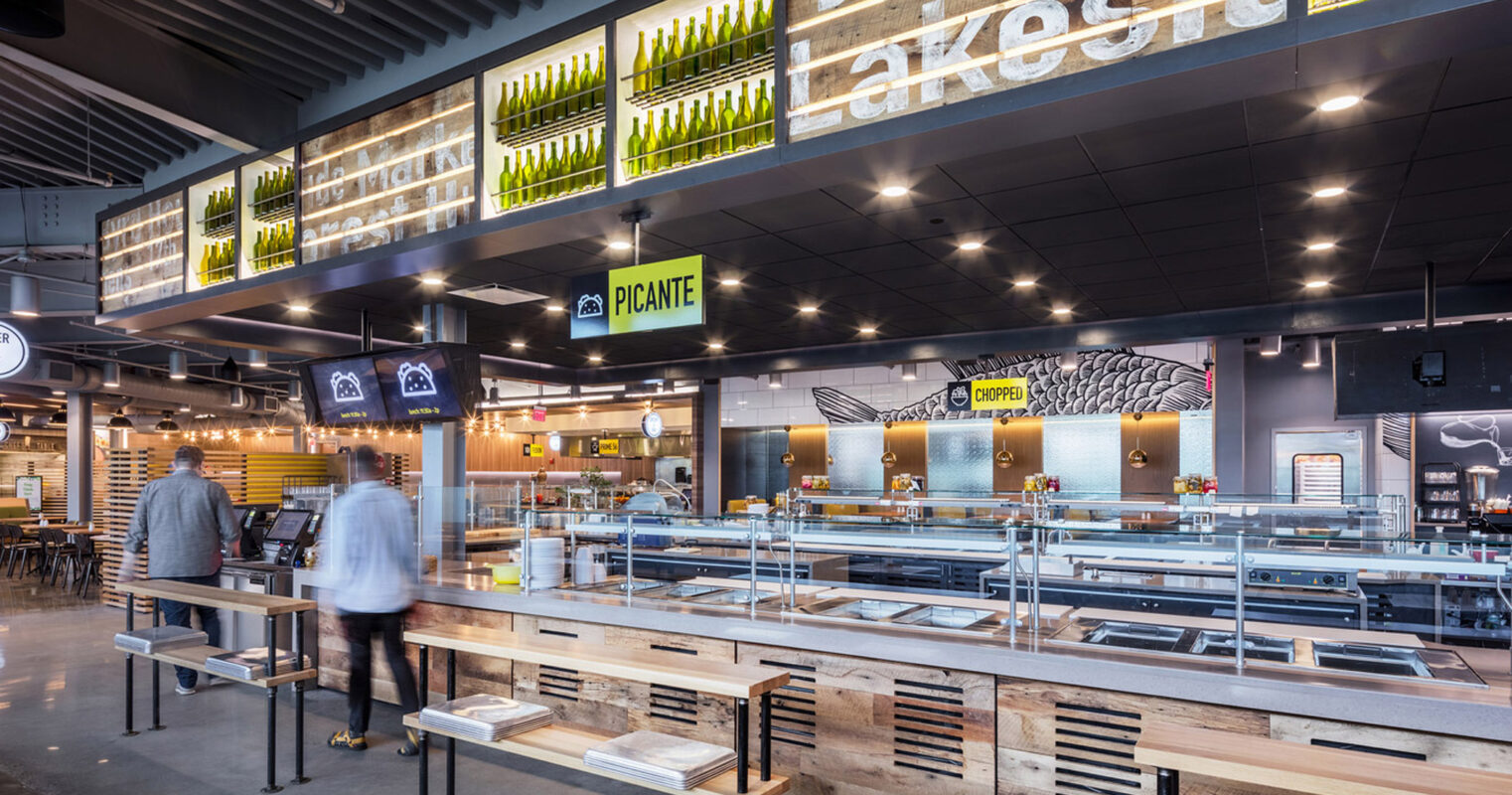 Modern commercial restaurant interior featuring an open kitchen design, with a harmonious mix of industrial ceiling elements and warm, natural wood finishes. Suspended green planters and ambient lighting complement the space's inviting, contemporary aesthetic.