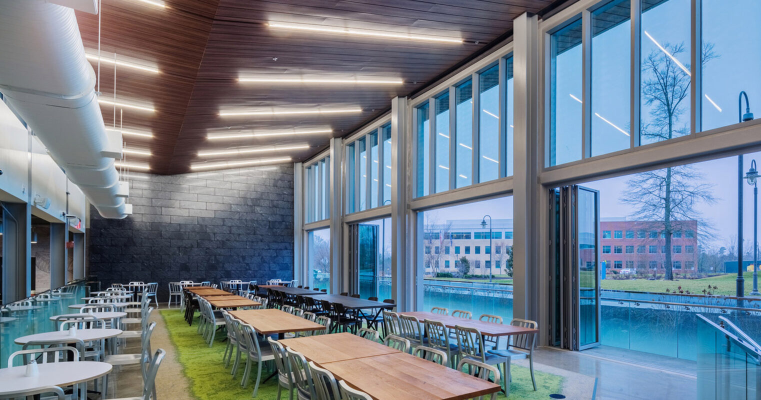 Spacious dining hall boasting high ceilings with wooden slats, wall-to-wall glazing providing ample natural light, and modern rectangular tables on a vibrant green carpet, complementing the exterior view of serene waters and architecture.