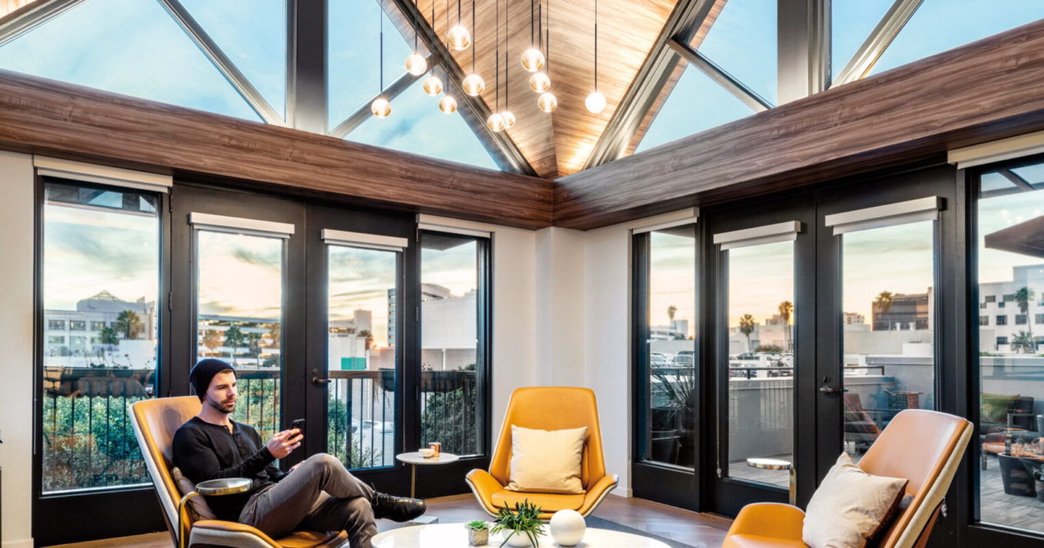 Vaulted ceiling with exposed wooden beams converges to a skylight, complemented by floor-to-ceiling glass walls. Modern furnishings include a central round table, mustard yellow armchairs, and black accents, with an angled black floor lamp adding contrast in a warm, natural-lit room. A person reclines, absorbed in a device.