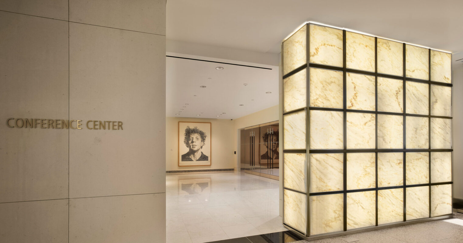 Elegantly lit translucent stone paneling flanks the entrance to a modern conference center, contrasting with a minimalistic matte wall bearing the center's signage. The warm glow adds ambience, harmonizing with the reflective flooring while highlighting adjacent artwork.