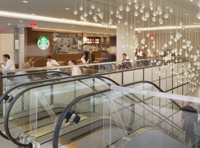 Modern commercial interior featuring an expansive, illuminated ceiling installation with cascading spherical lights over a dual staircase. Reflective surfaces and minimalist furniture provide a sleek, contemporary ambiance, complemented by a coffee shop counter on the upper level.