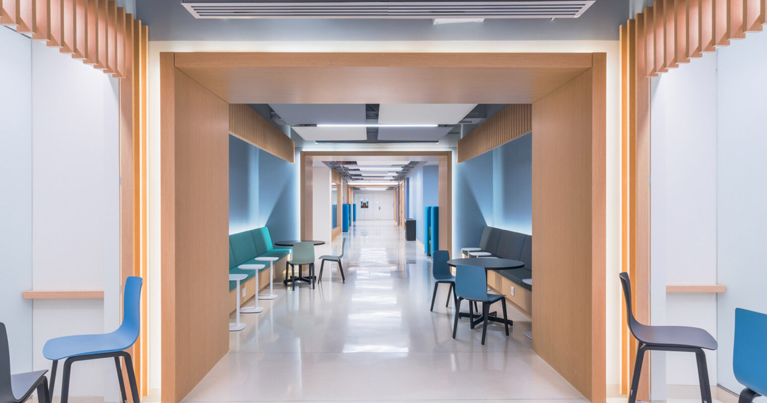 Modern corridor with clean lines, featuring wood-accented alcoves, teal and blue furnishing, and efficient LED lighting guiding the way. The design merges functionality with comforting aesthetics to ease navigation and enhance patient comfort.