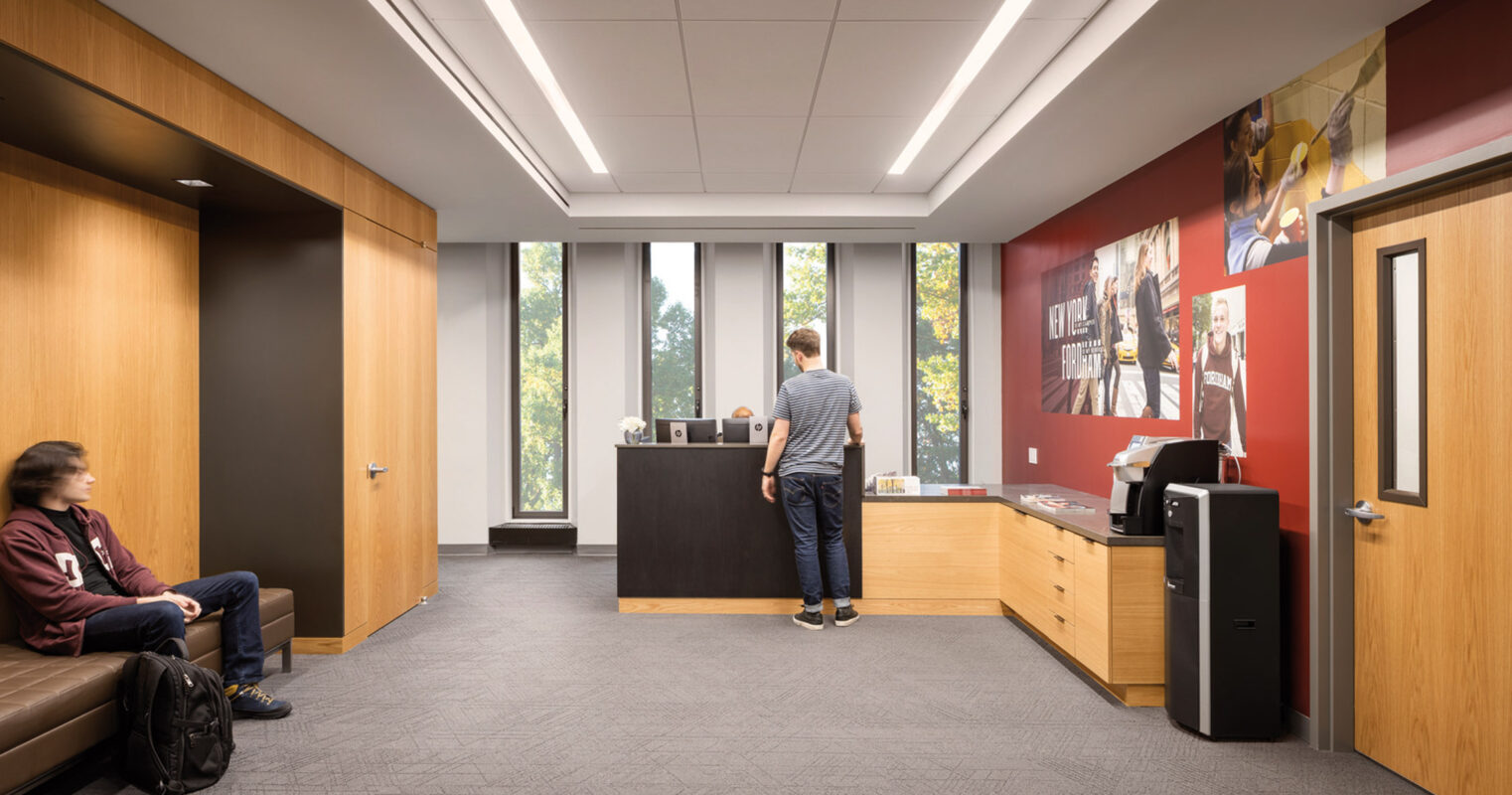Modern office waiting area with warm wood paneling, recessed linear lighting, and floor-to-ceiling windows offering a view of greenery. Two individuals calmly wait, one seated and another standing by the reception desk.