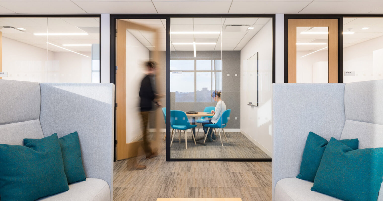 Modern office space featuring a glass-walled meeting room with a wooden table and chairs, flanked by cozy grey couches with vibrant blue cushions, and a light wood coffee table. A sense of activity is captured by a blurred figure passing by.