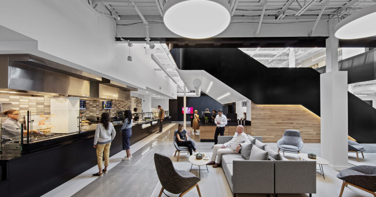 Spacious office breakroom with modern design elements, featuring a sleek black kitchenette, round overhead lighting, and a mix of casual seating arrangements with organic color accents.