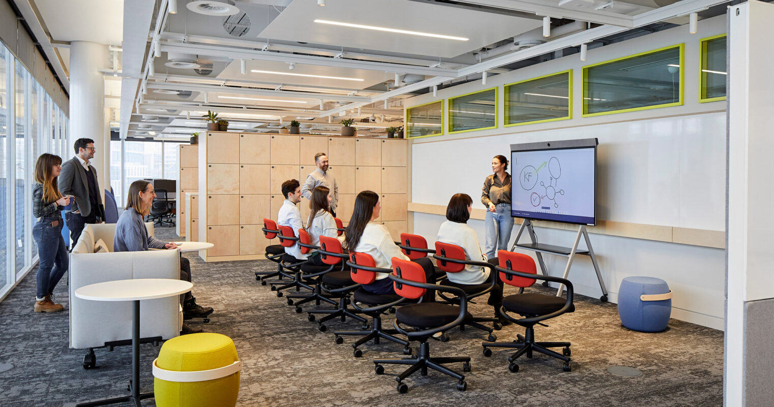 Modern office interior featuring a collaborative workspace with amphitheater-style seating, vibrant color accents, and a digital presentation display. Wooden slat partitions offer structural detail and acoustic benefits while natural light enhances the welcoming atmosphere.