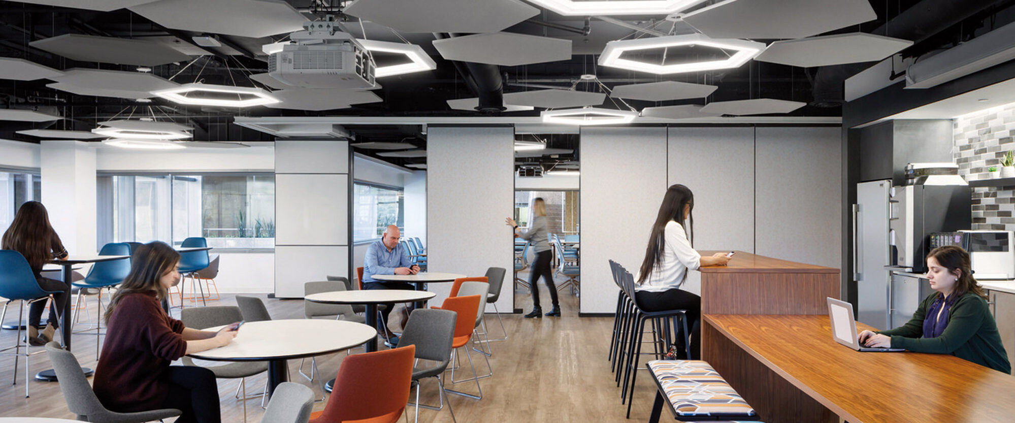 Modern office cafeteria featuring an eclectic mix of hexagonal and circular suspended lighting fixtures. Geometric ceiling panels complement the angular motif. Varied seating options including communal tables and private booths cater to diverse preferences, each delineated by contrasting color schemes and textures.