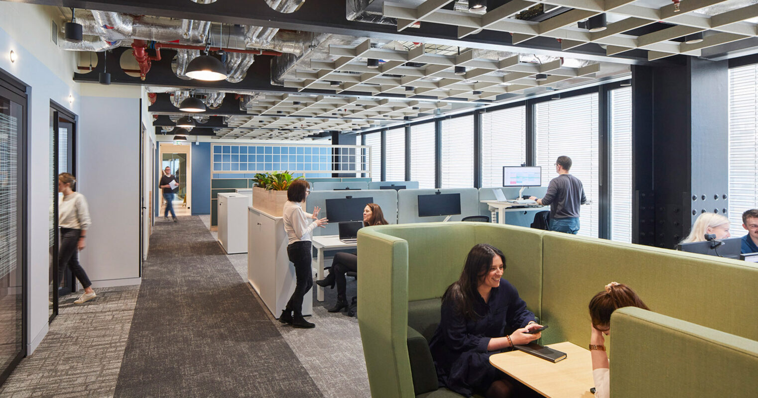 Contemporary office space featuring an open-plan layout with individual workstations. Green upholstered privacy couches offer informal seating areas. Overhead, a geometric ceiling with exposed ductwork juxtaposes the organized, clean lines of the modular, functional furniture below. Natural light enhances the productivity-focused design.