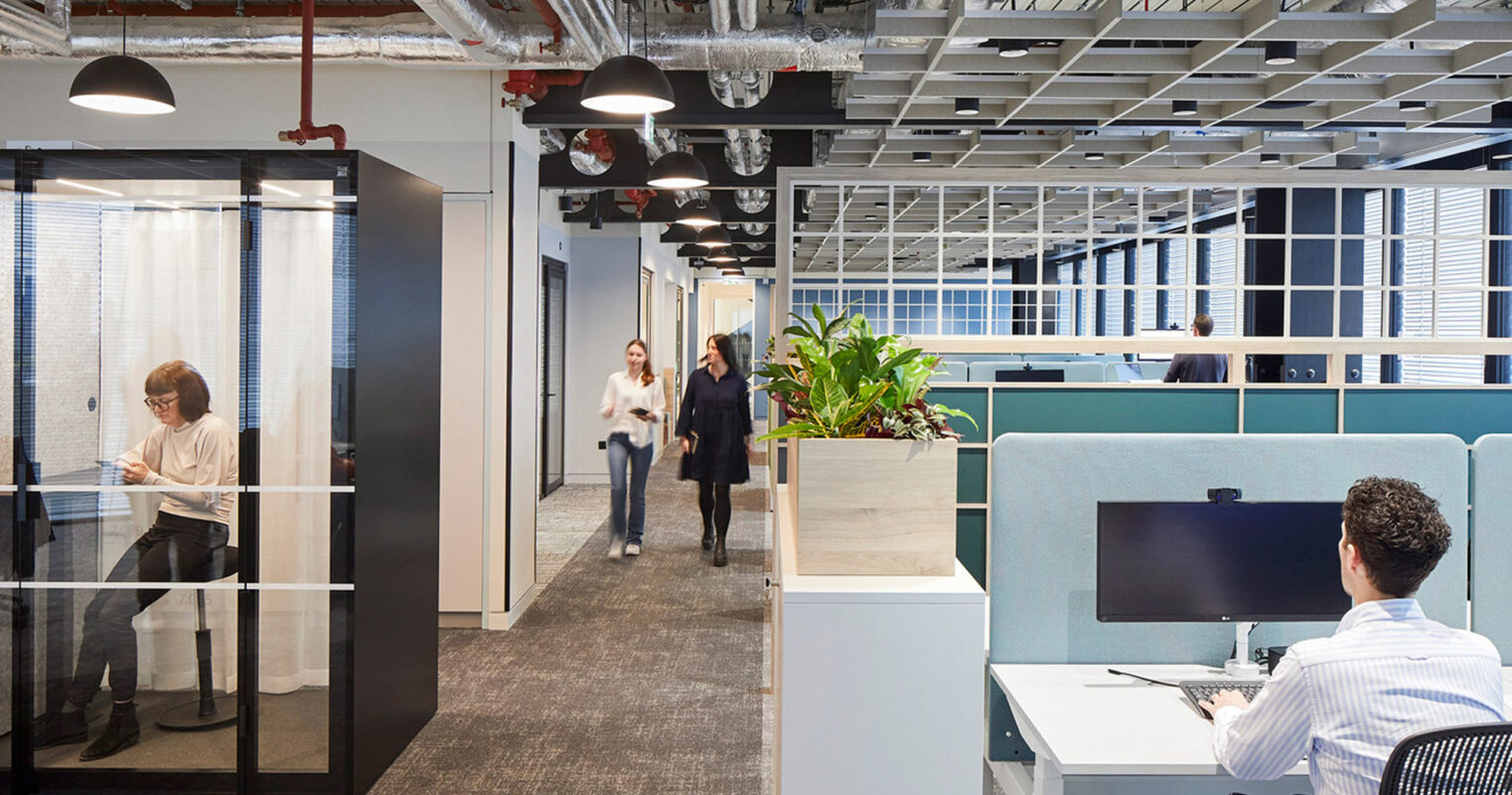 Modern office interior featuring an open floor plan with individual workstations, glass-walled meeting rooms, exposed ceiling beams, and vibrant blue privacy booths. Natural light and pedestrian movement evoke a dynamic work environment.