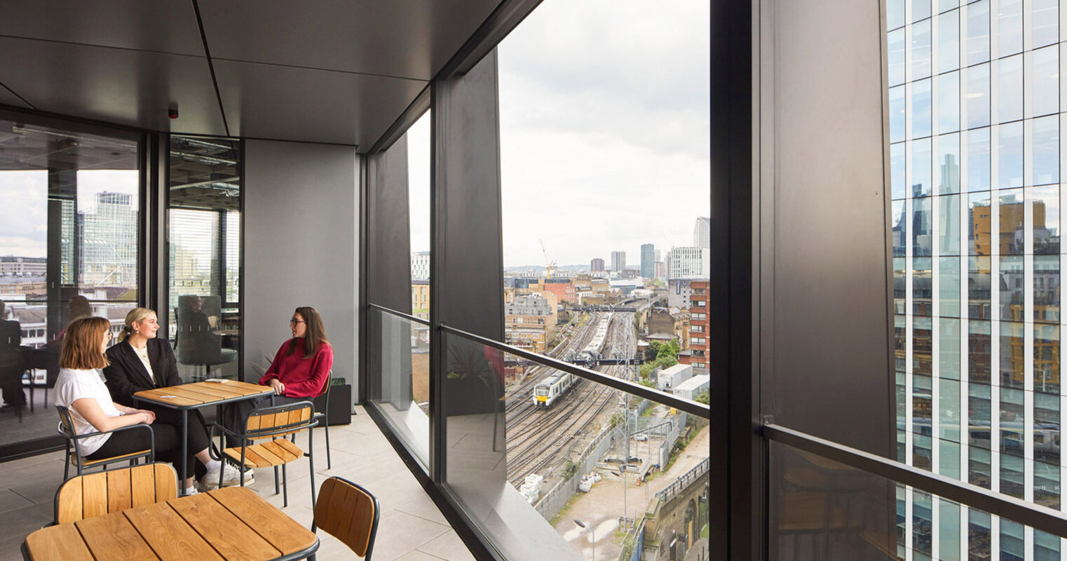 Modern office space with floor-to-ceiling windows offering sweeping city views, furnished with minimalist wooden desks and elegantly simple seating for a collaborative meeting. The room's clean lines and abundant natural light epitomize contemporary corporate design.