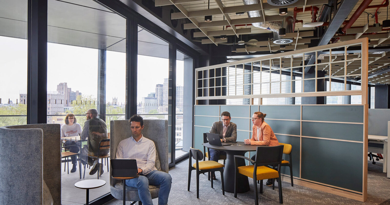 Modern open-plan office space with glass partitions, featuring a mix of casual seating and collaborative work areas. Subtle neutral tones are accented with pops of yellow, while natural light floods the room creating a bright and inviting atmosphere.