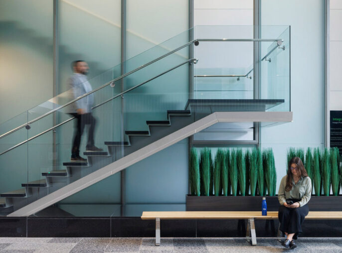 Contemporary office lobby with a floating staircase featuring glass balustrades and sleek metal handrails. Transparent partition walls create a spacious feel, while a person ascends the stairs and another sits on a minimalist bench with lush greenery underneath, underscoring a biophilic design approach.