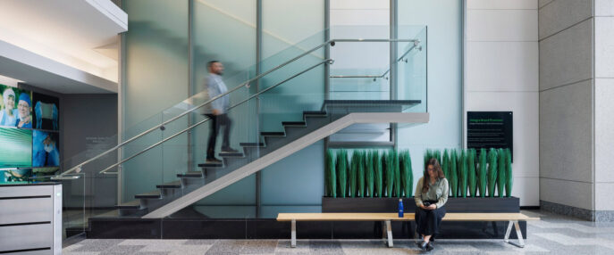 Contemporary office lobby with a floating staircase featuring glass balustrades and sleek metal handrails. Transparent partition walls create a spacious feel, while a person ascends the stairs and another sits on a minimalist bench with lush greenery underneath, underscoring a biophilic design approach.