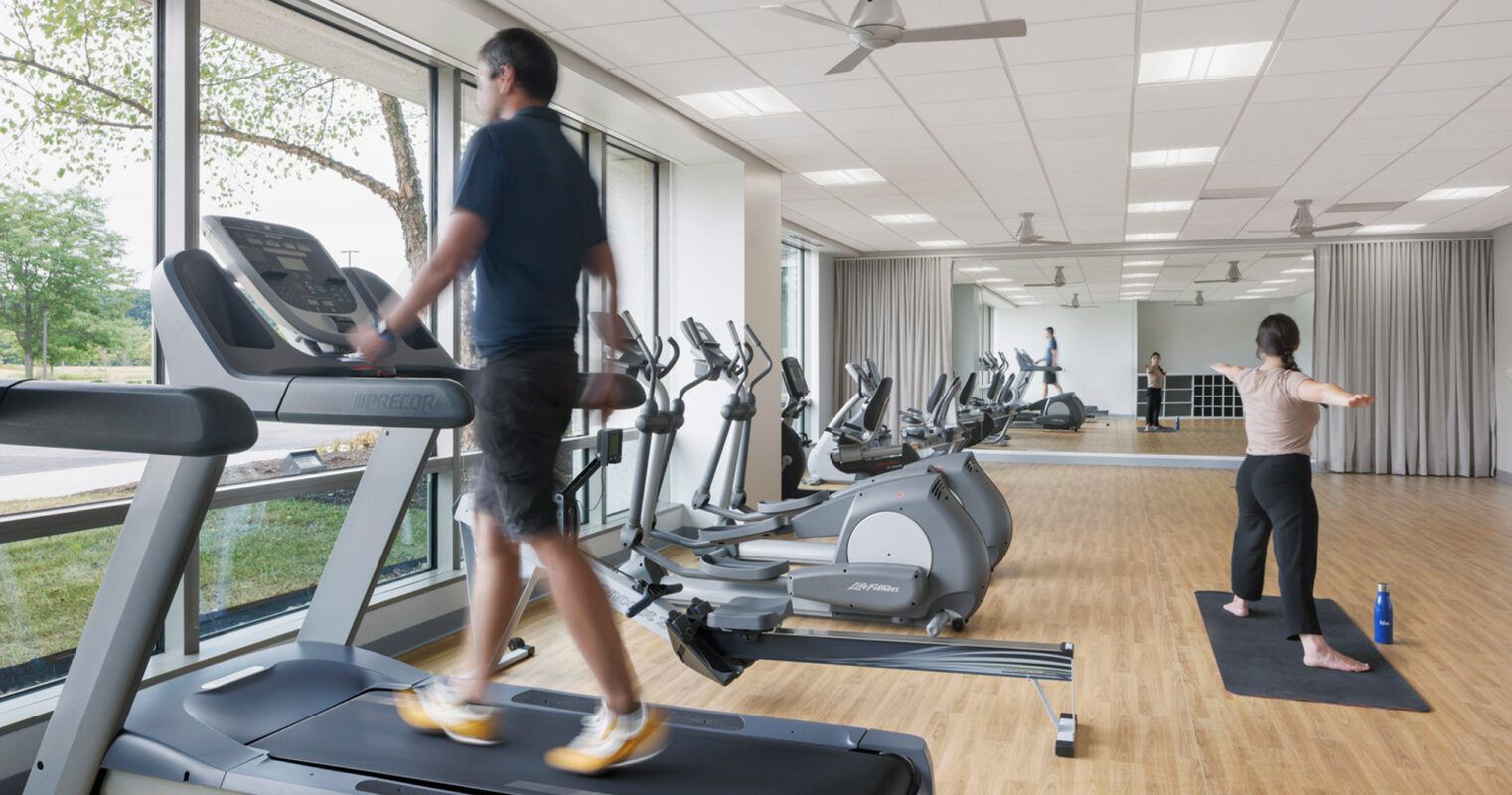 Bright and airy fitness center with front-facing treadmills, stationary bikes, and elliptical machines; ample daylight streams in through large windows. Two individuals engage in exercise, one on a treadmill, another practicing yoga, highlighting the facility's versatility for various workout routines.