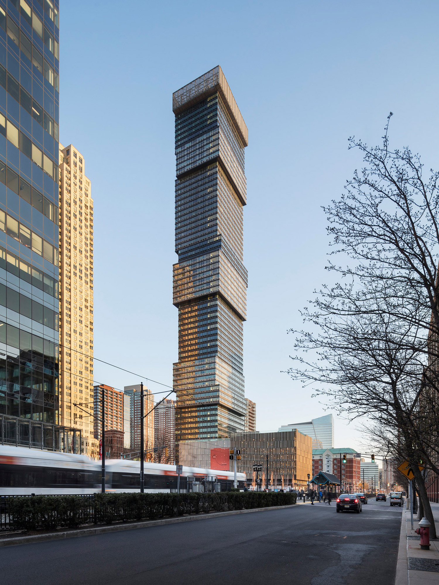 Modern skyscraper with a dynamic, staggered façade showcasing a mix of glass and metallic cladding, reflecting the urban surrounding and sky. The tower's structure presents a vertical juxtaposition that plays on light and shadow, emphasizing the innovative architectural approach within a bustling city environment.