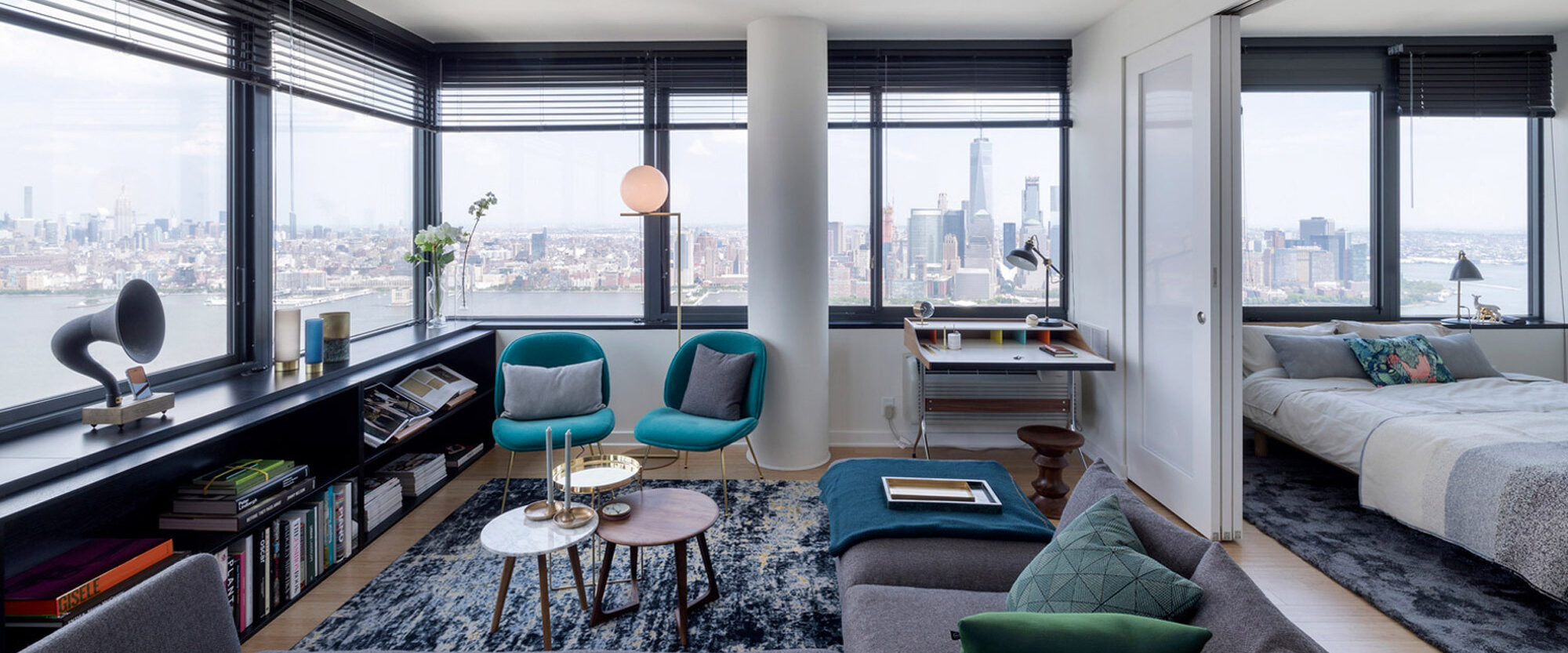 Open-plan living space with floor-to-ceiling windows offering a panoramic city view. Mid-century modern influences seen in teal armchairs and geometric coffee tables. Neutral tones balance the vibrant furniture, and strategic lighting enhances the room's sleek, urban ambiance.
