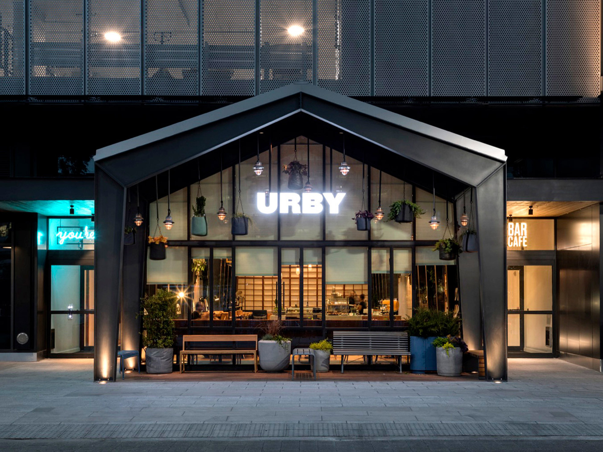 Modern urban cafe exterior with symmetrical design, featuring warm interior lighting, hanging plants, and a welcoming bench under a canopy, flanked by potted greenery.