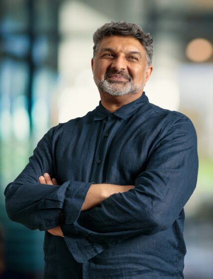 The photograph features a man with salt-and-pepper hair and a beard, giving a slight, confident smile, wearing a dark collared shirt with the sleeves rolled up to his elbows. He stands with his arms crossed in front of him against a light and airy office backdrop with out-of-focus lights, suggesting a professional environment.