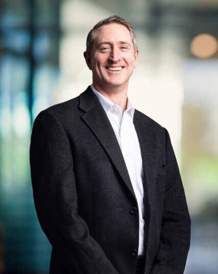 The image shows a man with a genuine smile, short-cropped hair, wearing a white shirt and a dark overcoat. He stands with his hands comfortably in his pockets, against a backdrop of blurred blue-toned lights that suggest a modern, professional setting.