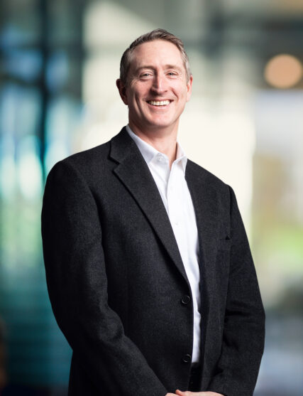 The image shows a man with a genuine smile, short-cropped hair, wearing a white shirt and a dark overcoat. He stands with his hands comfortably in his pockets, against a backdrop of blurred blue-toned lights that suggest a modern, professional setting.