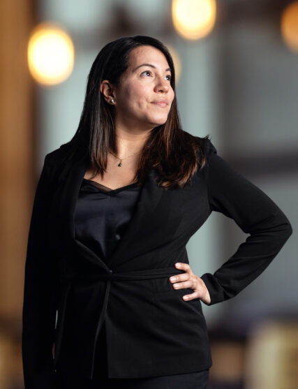 The image depicts a woman with long dark hair and a contemplative expression, dressed in a black blazer and top, with her hand on her hip. The background features warm, circular bokeh lights, which contribute to a sophisticated atmosphere likely within an office setting. The image depicts a woman with long dark hair and a contemplative expression, dressed in a black blazer and top, with her hand on her hip. The background features warm, circular bokeh lights, which contribute to a sophisticated atmosphere likely within an office setting. The image depicts a woman with long dark hair and a contemplative expression, dressed in a black blazer and top, with her hand on her hip. The background features warm, circular bokeh lights, which contribute to a sophisticated atmosphere likely within an office setting.