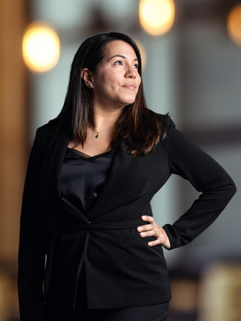 The image depicts a woman with long dark hair and a contemplative expression, dressed in a black blazer and top, with her hand on her hip. The background features warm, circular bokeh lights, which contribute to a sophisticated atmosphere likely within an office setting. The image depicts a woman with long dark hair and a contemplative expression, dressed in a black blazer and top, with her hand on her hip. The background features warm, circular bokeh lights, which contribute to a sophisticated atmosphere likely within an office setting. The image depicts a woman with long dark hair and a contemplative expression, dressed in a black blazer and top, with her hand on her hip. The background features warm, circular bokeh lights, which contribute to a sophisticated atmosphere likely within an office setting.