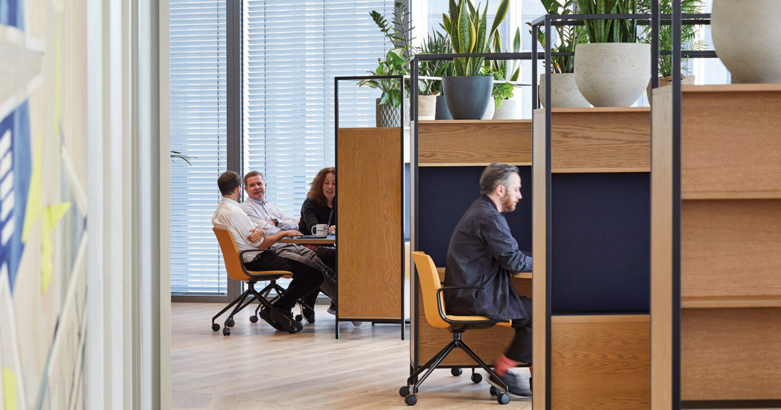 Modern office space with natural light, featuring wood-accented bookshelves doubling as dividers. Open workstations and a collaborative area with green planters cultivate a warm, productive ambiance.