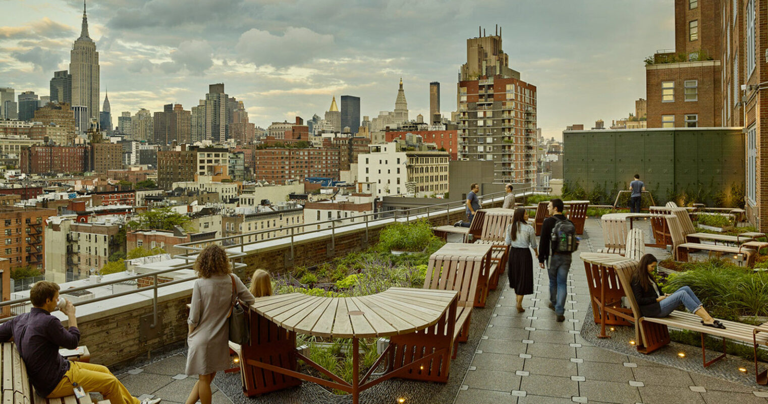 Rooftop terrace with wooden seating and ample greenery, featuring urban skyline views, under a dusk sky. The space balances modern and rustic elements, integrating with the cityscape.