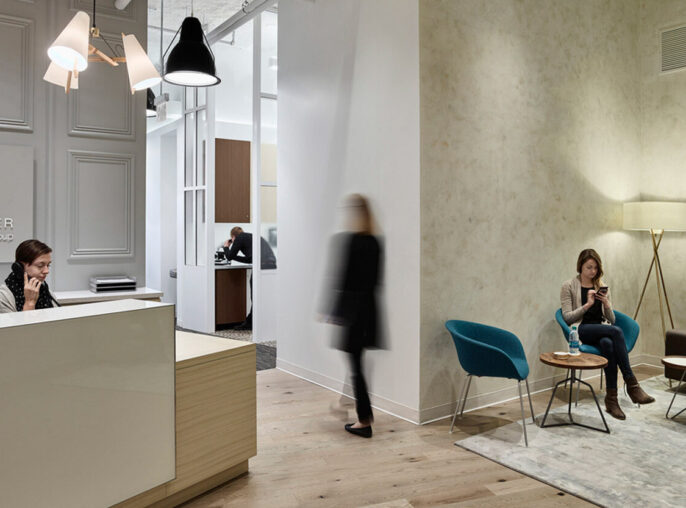 Elegant wellness center lobby featuring contrasting textures with smooth, ornate wall paneling and rustic, exposed concrete. A minimalist reception desk pairs with modern, black lighting fixtures, while patrons relax in stylish armchairs on a muted area rug over weathered hardwood flooring.