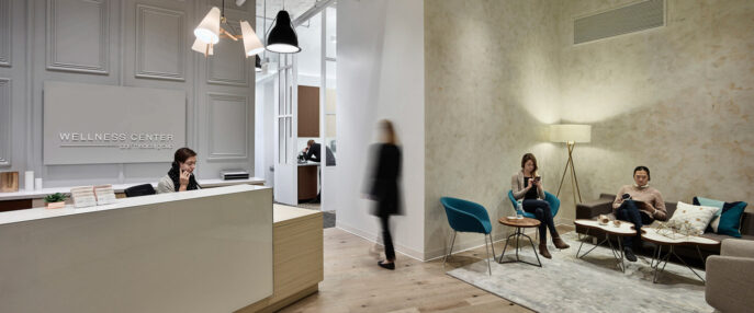 Elegant wellness center lobby featuring contrasting textures with smooth, ornate wall paneling and rustic, exposed concrete. A minimalist reception desk pairs with modern, black lighting fixtures, while patrons relax in stylish armchairs on a muted area rug over weathered hardwood flooring.