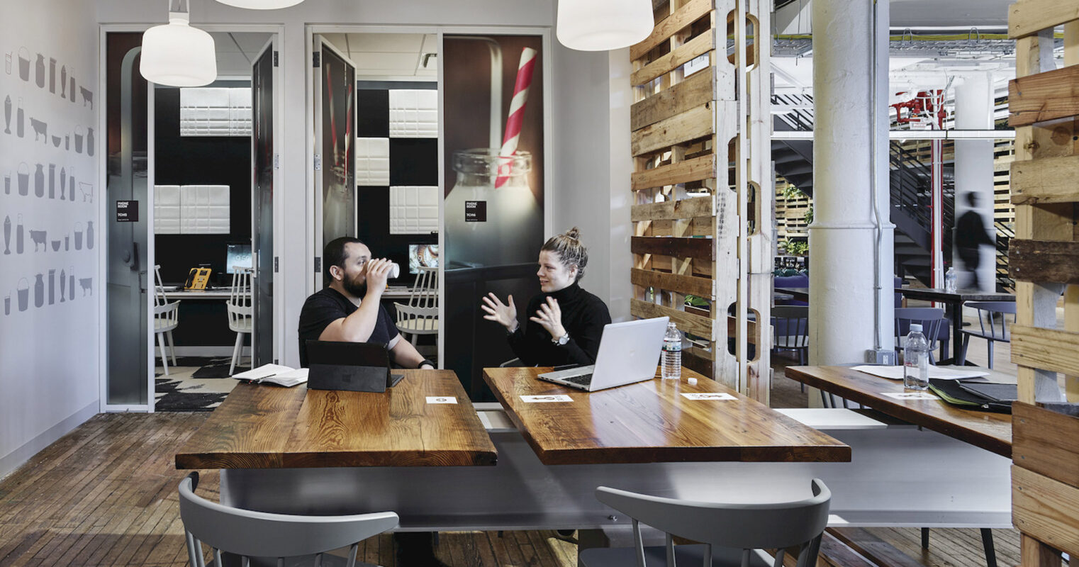 Modern open-concept office space with exposed ceiling and natural wood accents, featuring pendant lighting above a communal table where two individuals engage in conversation. The design incorporates a balance of industrial and organic elements, creating a dynamic, collaborative environment.