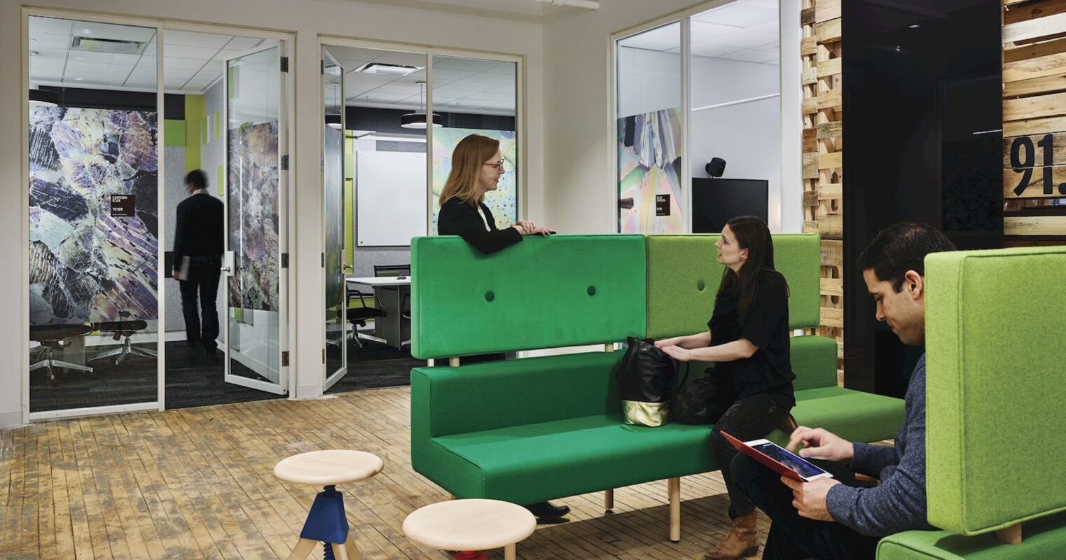 Modern office space featuring bright green upholstered seating with tall backrests for privacy. Wood flooring and accent wall add warmth, while the use of glass partitions maintains transparency. Unique chandelier lighting complements abstract wall art, enhancing the space's creative ambiance.