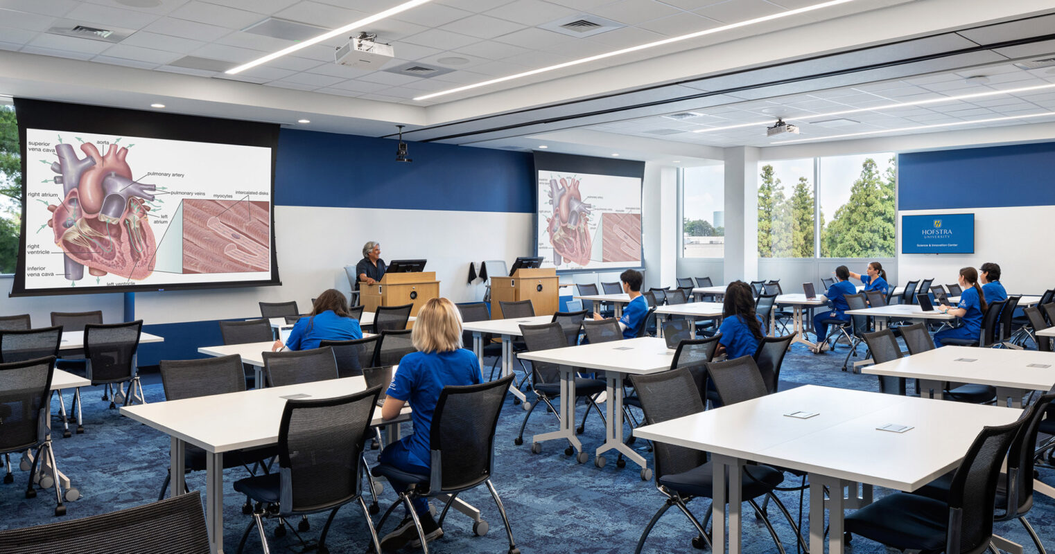 Spacious classroom featuring modern movable tables and chairs with a blue and white color scheme. The front wall displays a large projection screen, while ample natural light enhances the learning environment.