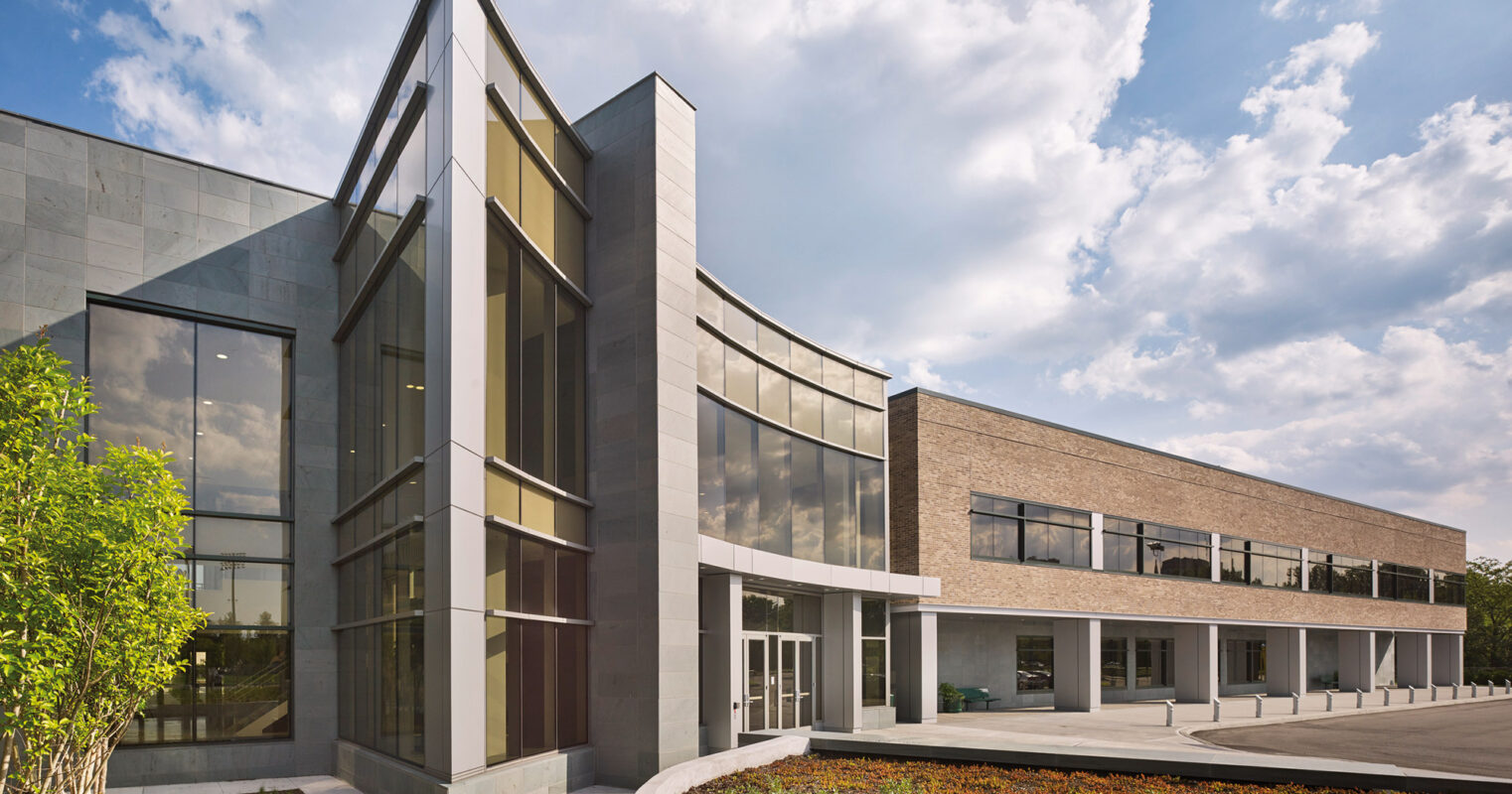 Modern architectural exterior featuring a juxtaposition of sleek, reflective glass and matte stone finishes. Angled rooflines and a mix of vertical windows create a dynamic facade, with landscaped greenery complementing the structure's geometric precision.